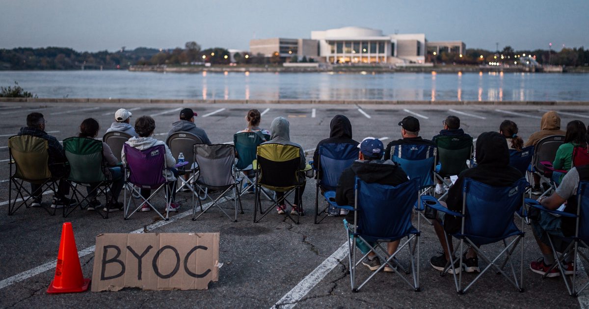 Wide shot at dusk on a riverside parking area beside the Potomac, several dozen people seen from behind sitting on folding camp chairs facing an empty stretch.
