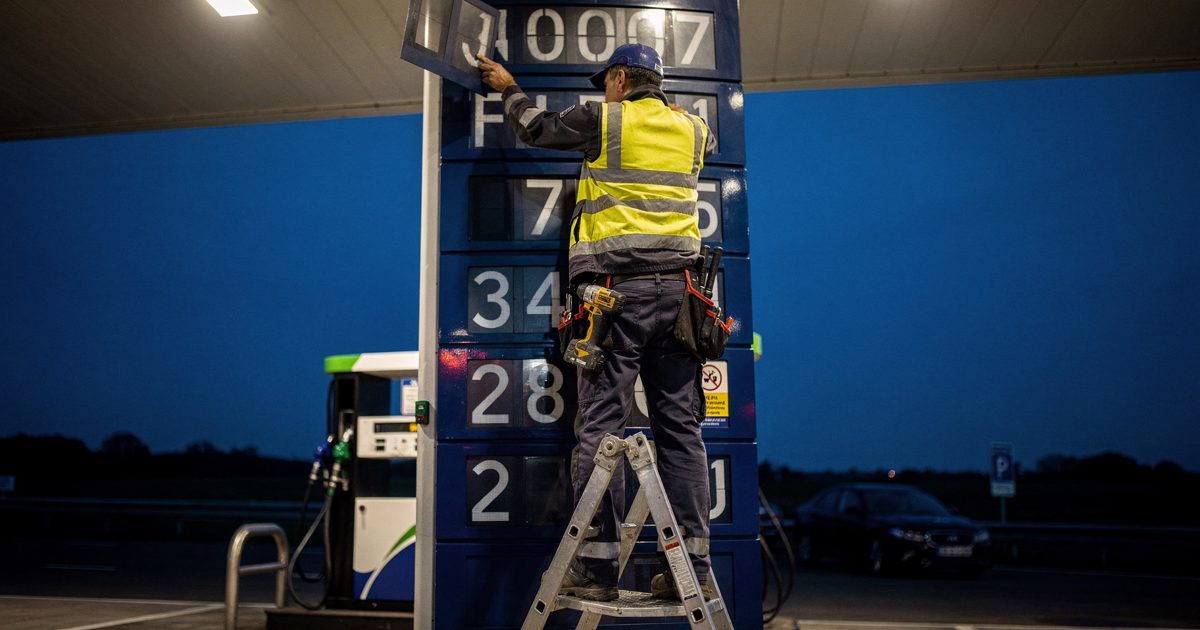 Deep blue dusk at a motorway petrol station forecourt, a maintenance worker seen from directly below on a stepladder with the ladder and canopy lights hiding.