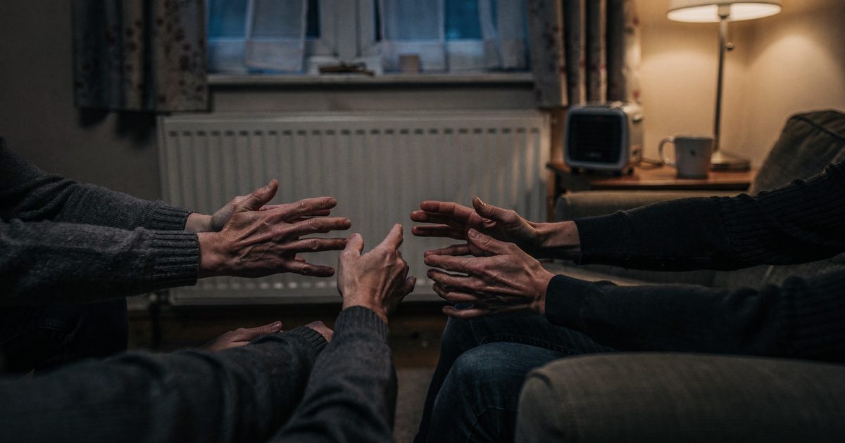 Close shot at knee height in a dim terraced-house living room on a winter evening lit by a single floor lamp, several pairs of hands from different people.