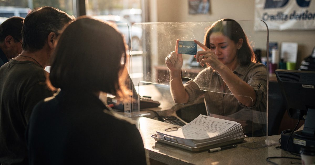 Medium shot from across a teller line inside a strip-mall credit union on Sunday afternoon, warm sun through the front doors, customers waiting with their.