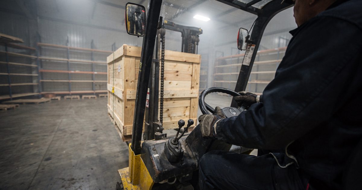 Under the harsh glare of floodlights at a predawn shipping terminal, a forklift driver's gloved hands grip the steering wheel while maneuvering a single crate.