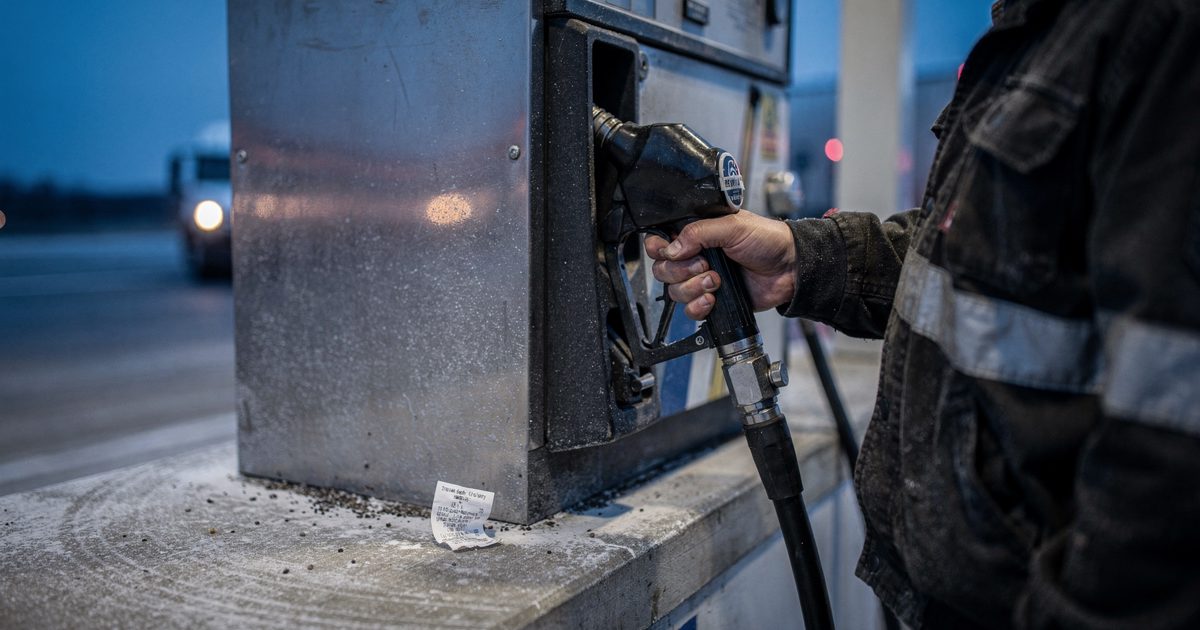 Close detail at a highway truck stop at blue pre-dawn light, a trucker cropped at the shoulder lifting a diesel nozzle from its pump holster, frost on the.