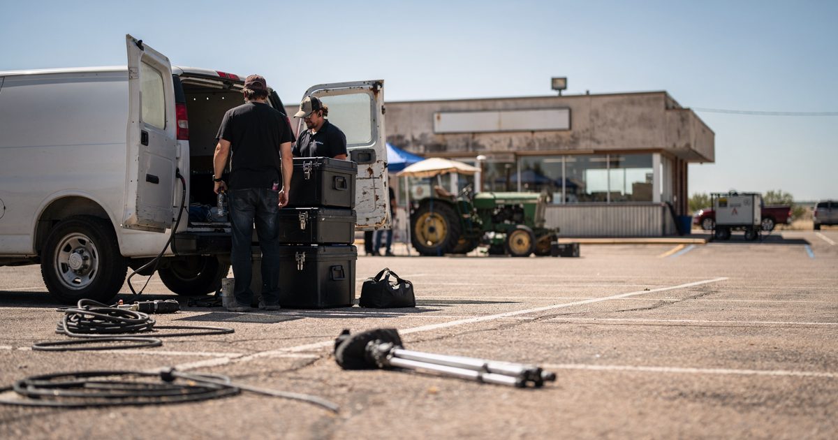 A sun-bleached Nebraska diner parking lot at high noon, where partially obscured production assistants unload heavy black equipment cases next to a weathered.