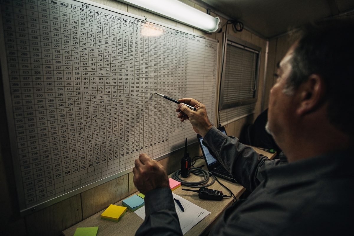 The dim, fluorescent-lit interior of a temporary production trailer, where a pair of hands points a pen at a sprawling wall calendar that ends in the year 2056.