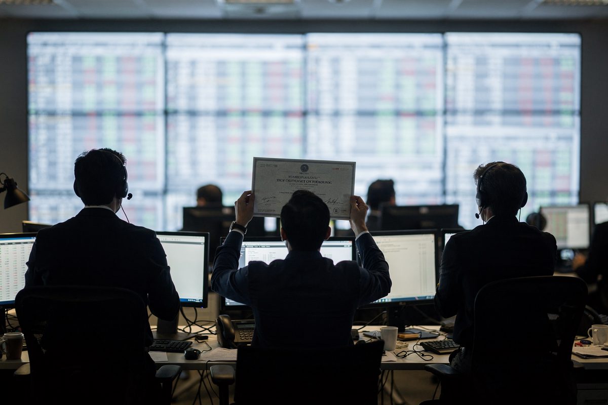 Medium shot inside a Rotterdam commodities trading floor at mid-morning, three traders reduced to silhouettes against a bright wall of price screens, one.