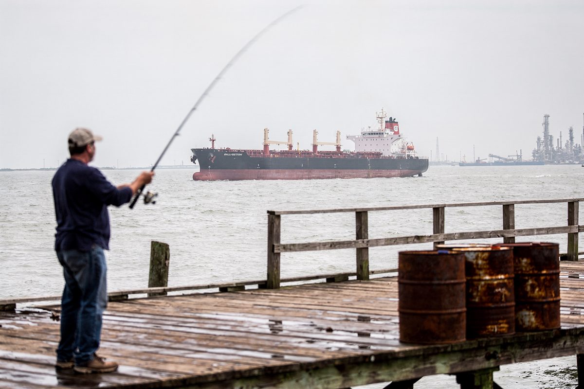 Wide shot at noon under a flat white sky from a Galveston Bay fishing pier, an angler blurred by motion in the foreground casting a line while a tanker rides.