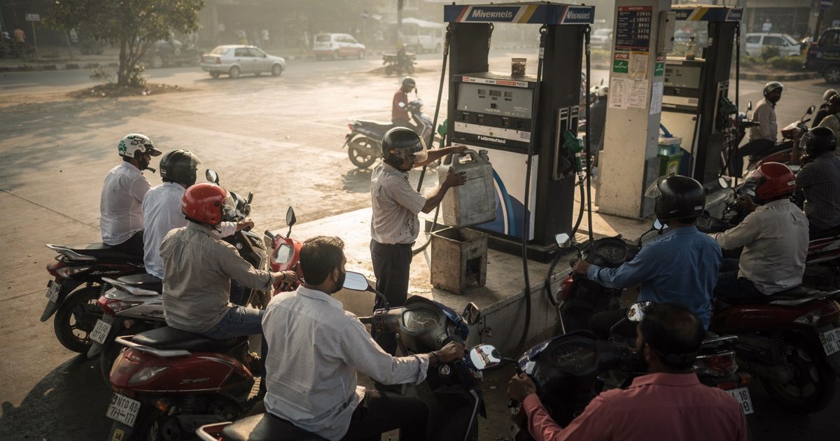 High-angle shot in pale early light over a New Delhi petrol-station forecourt, motorists on scooters clustering around the pumps while an attendant in a work.