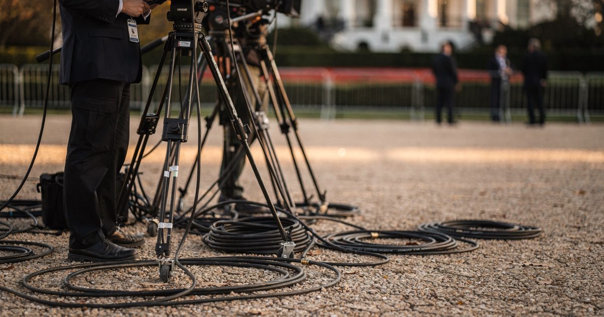 Mid-shot at golden hour on the gravel camera positions of the White House North Lawn, television crew members seen from the waist down uncoiling black cable.