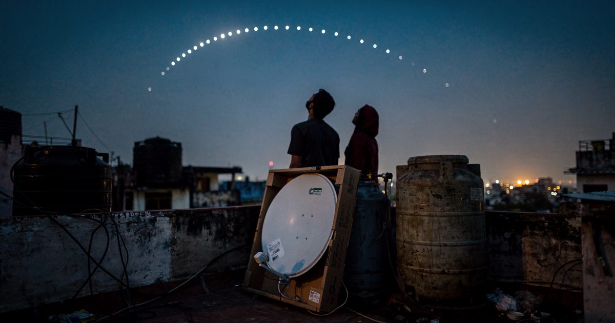 Low-angle shot from a Kolkata apartment rooftop in the deep blue hour before dawn, two figures seen only as silhouettes against the sky craning their necks.