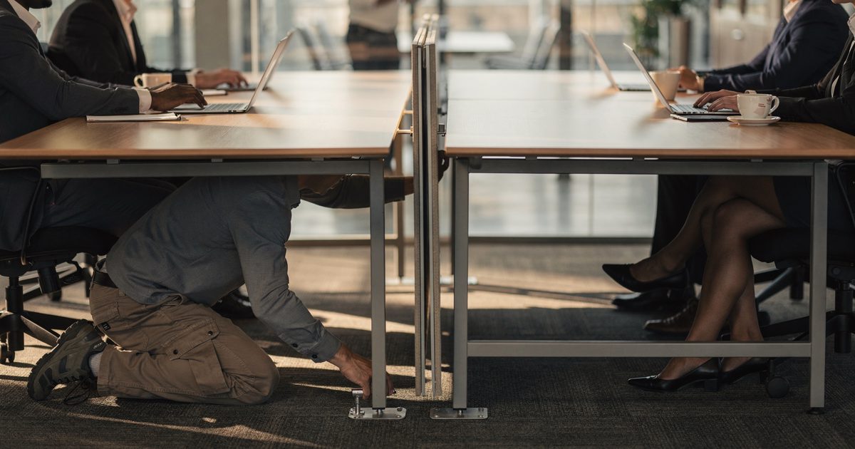 Waist-down shot in a glass-walled corporate meeting room in warm late-morning sun, a facilities worker in work trousers kneeling to unbolt a folding partition.