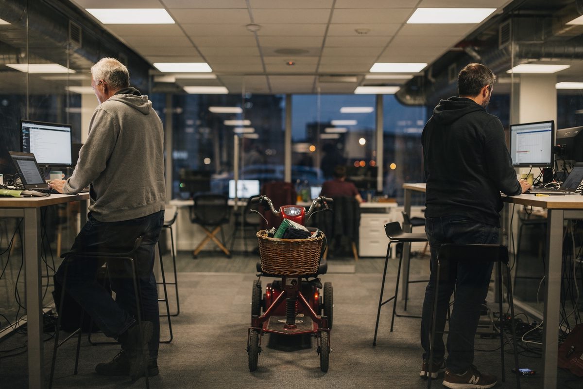 Wide dusk shot inside a glass-walled Shoreditch co-working floor under harsh white LED panels, two figures in hoodies seen from behind at standing desks while.