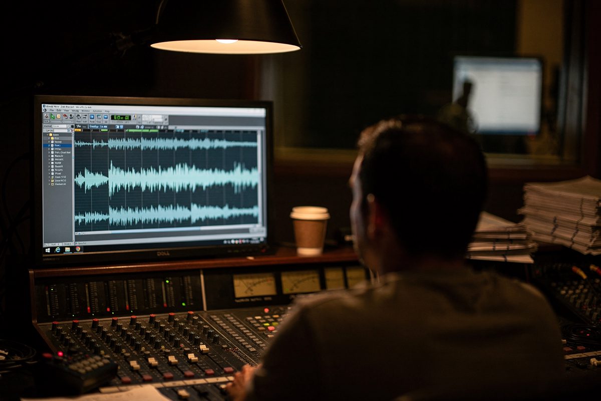 Under the glow of a single practical lamp inside a hushed dubbing stage, the capped head and shoulders of a mixer occupy the lower frame as a waveform scrolls.