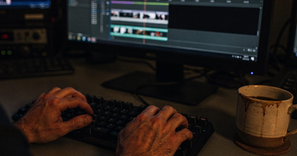 Close-up of hands in a dim editing suite scrubbing a timeline that ends abruptly at a single black frame, monitor glow on knuckles, an untouched mug of cold.