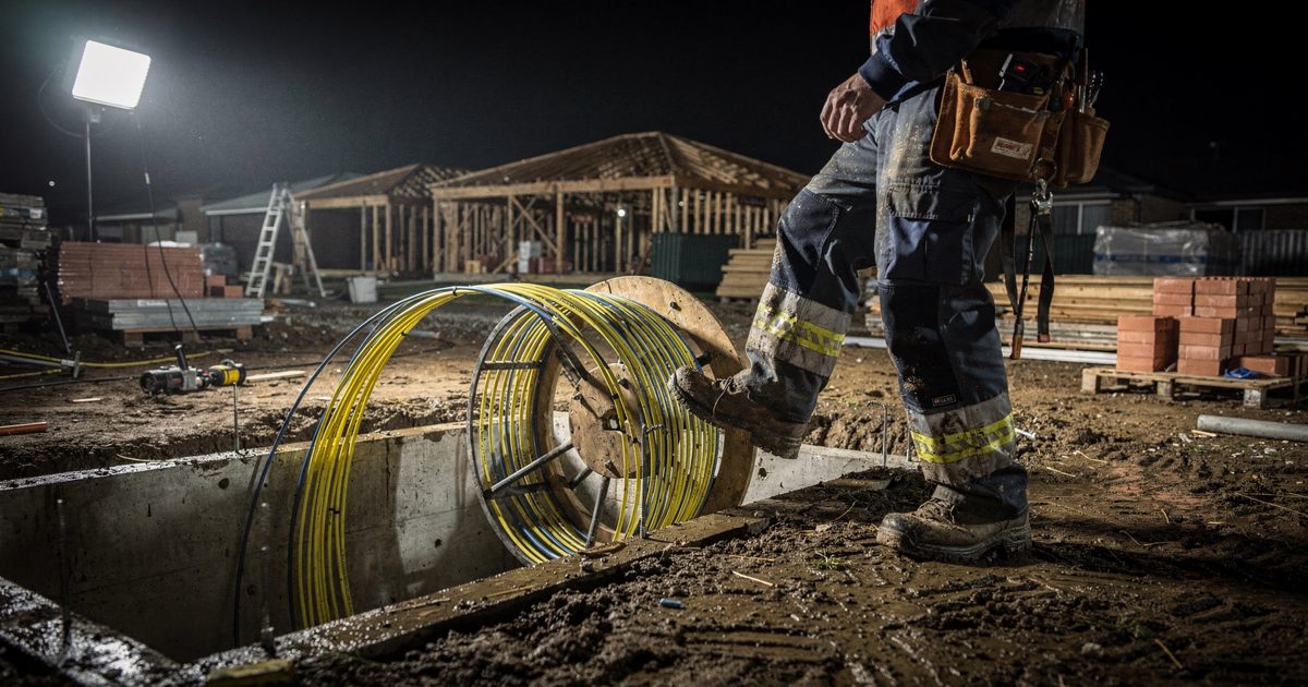Harsh halogen floodlights illuminating a muddy residential building site at midnight, a contractor framed from the waist down kicking a spool of fiber-optic.
