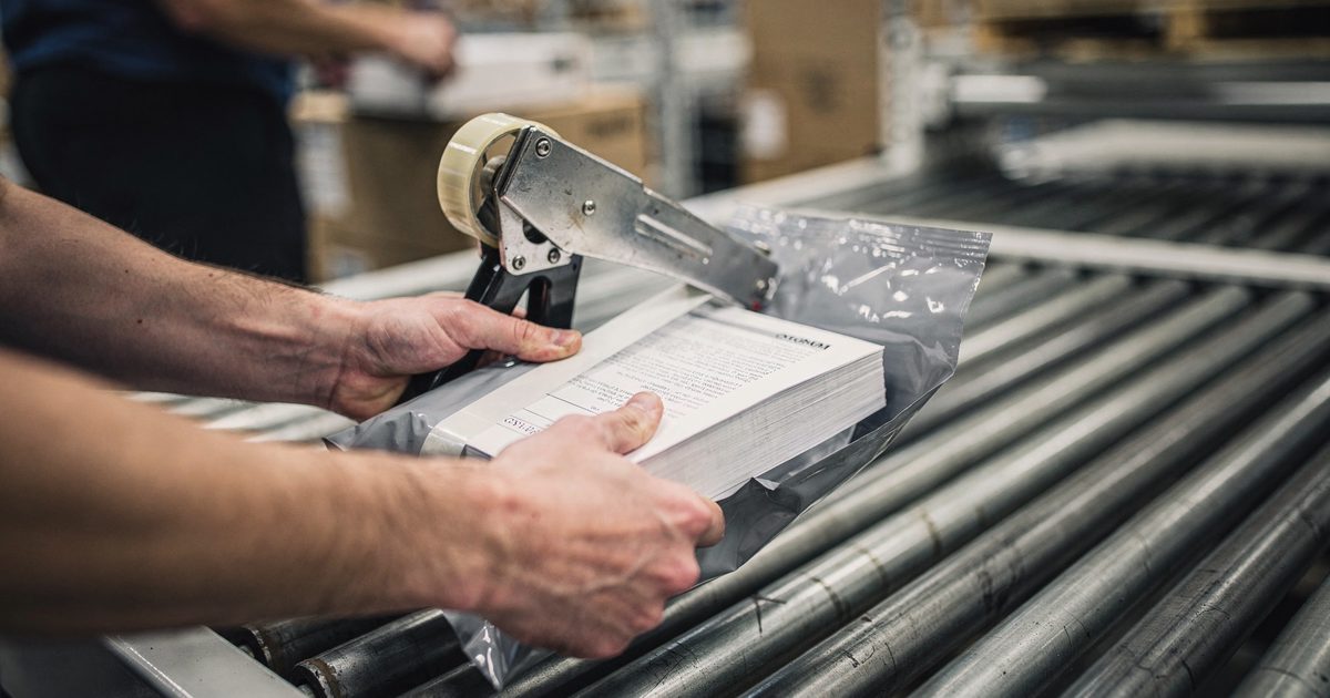 Close-up of hands in a fluorescent-lit fulfilment warehouse sealing a grey poly mailer stuffed with a thick bound document, packing tape gun mid-pull, conveyor.