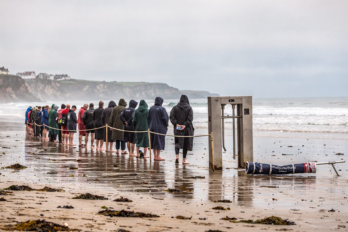 Wide morning shot of a Cornish beach at low tide where swimmers in dryrobes queue past a rope barrier toward the surf, a turnstile standing incongruously on.