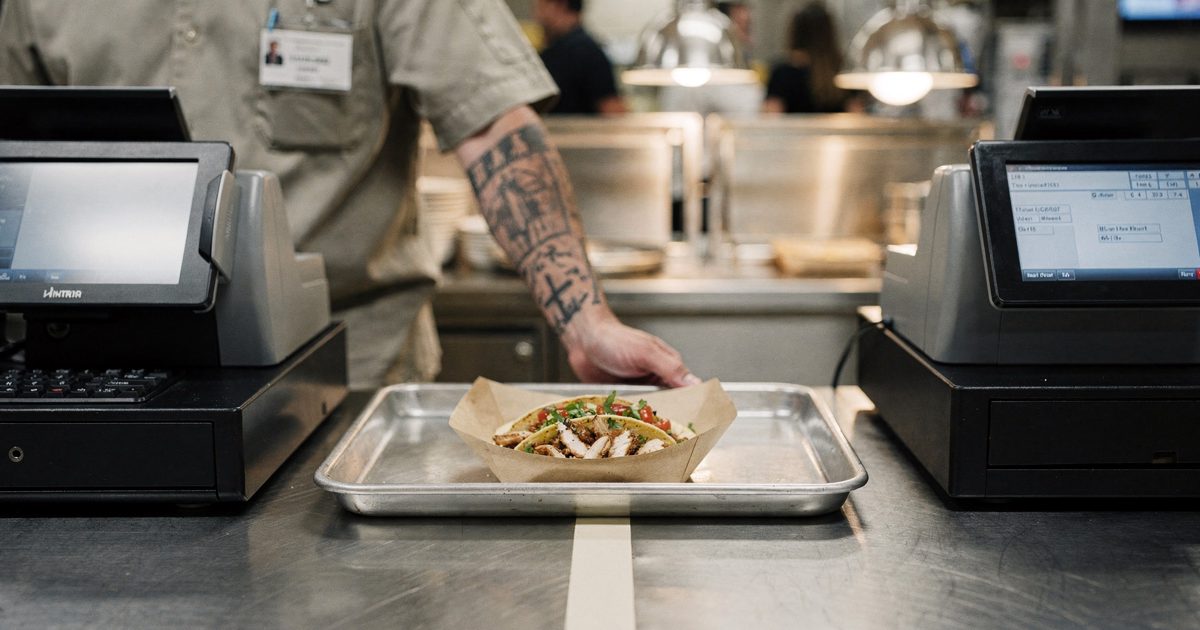 Close detail at bright midday inside a studio commissary, a single lunch tray on a steel counter holding one chicken taco set precisely between two cash.