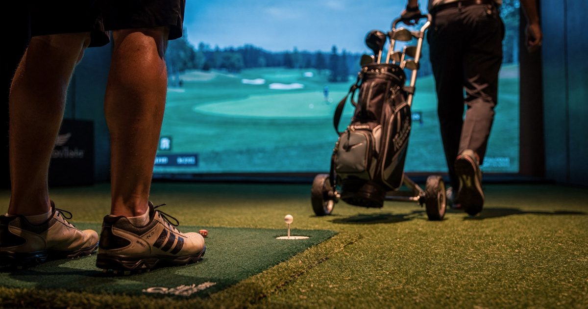 Low-angle shot from the turf of an indoor golf arena hitting bay at evening, lit by the cool blue glow of a giant simulator screen, an equipment staffer seen.
