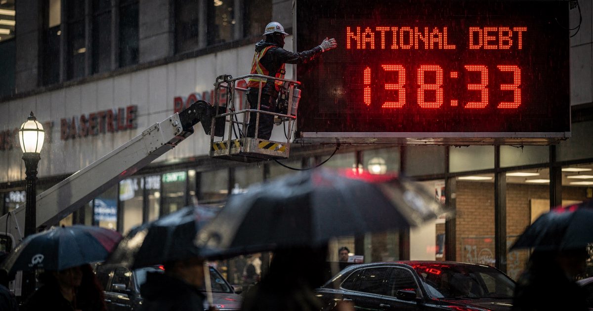 Night shot in light rain of the towering national debt clock above a Manhattan sidewalk, a maintenance worker in a raised cherry picker reaching toward the.