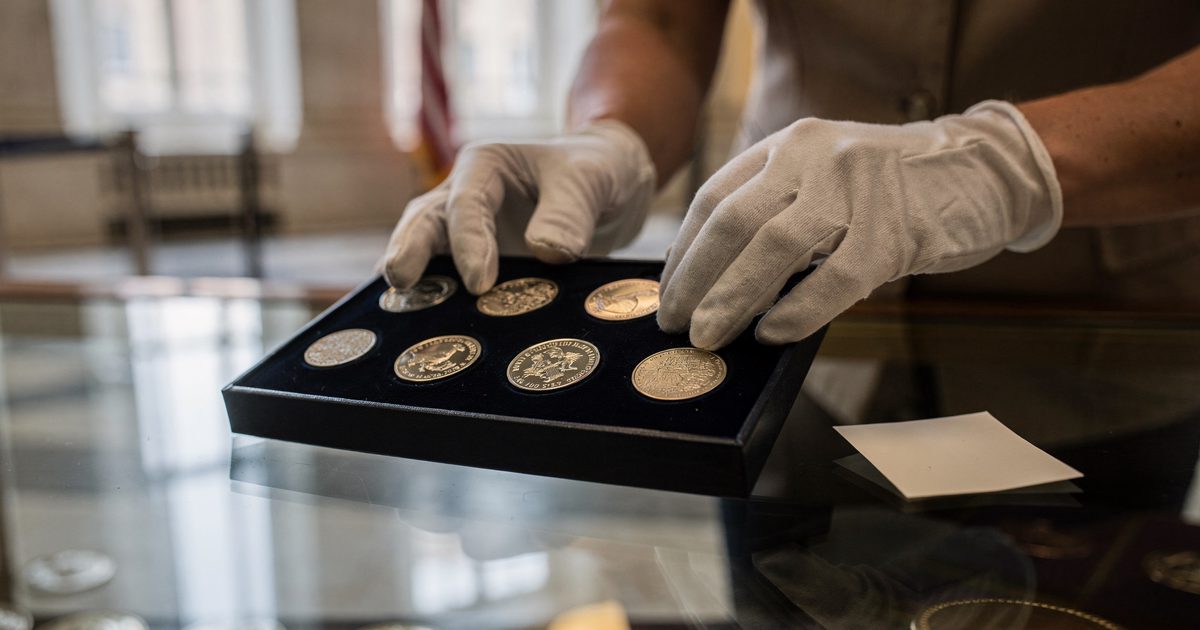 Close-up of gloved hands arranging a row of commemorative coins in a felt-lined display tray on a glass counter, early morning light through a government.