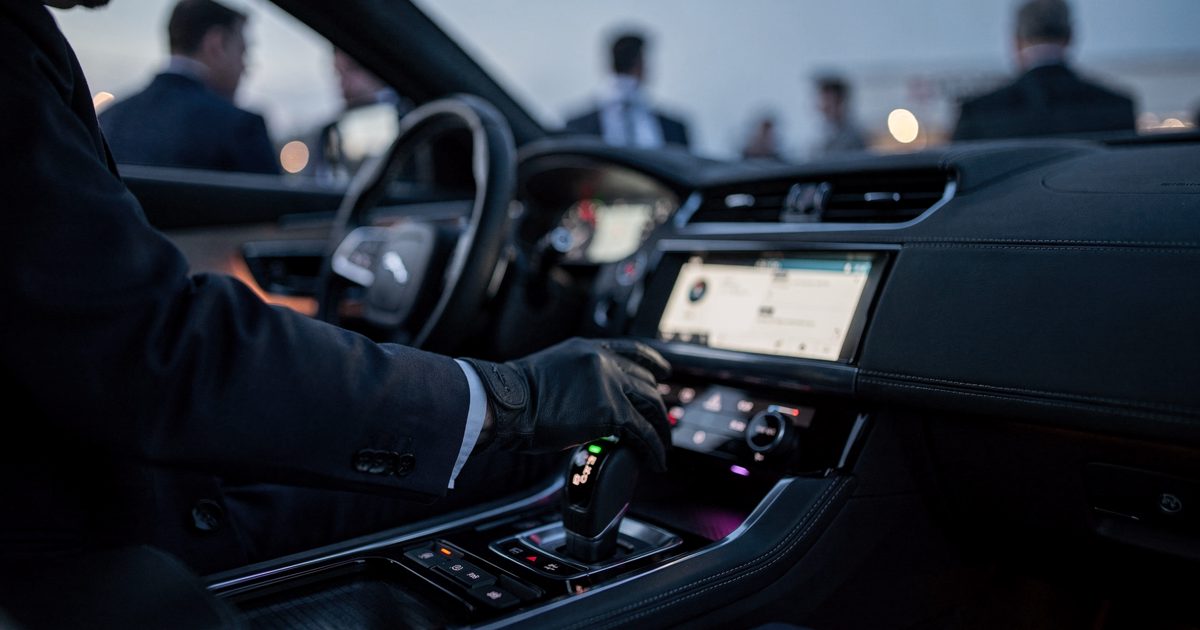 Close detail at blue-hour dusk on a car-maker's outdoor launch forecourt, a gloved hand of a staff member cropped at the forearm resting on the gear selector.
