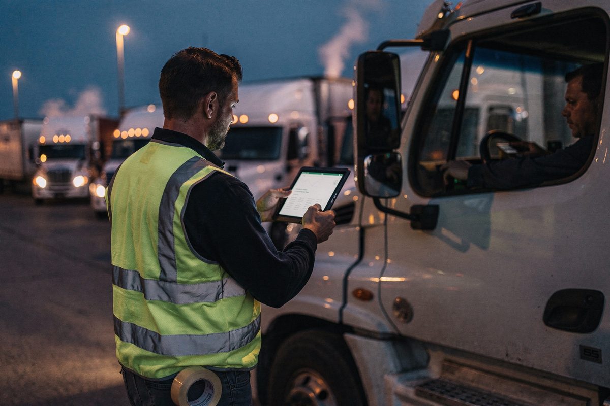 At dusk in a plaza of idling semis along the U.S.-Canada line, a worker seen from behind in a high-vis vest lifts a tablet toward a cab window with a roll of.