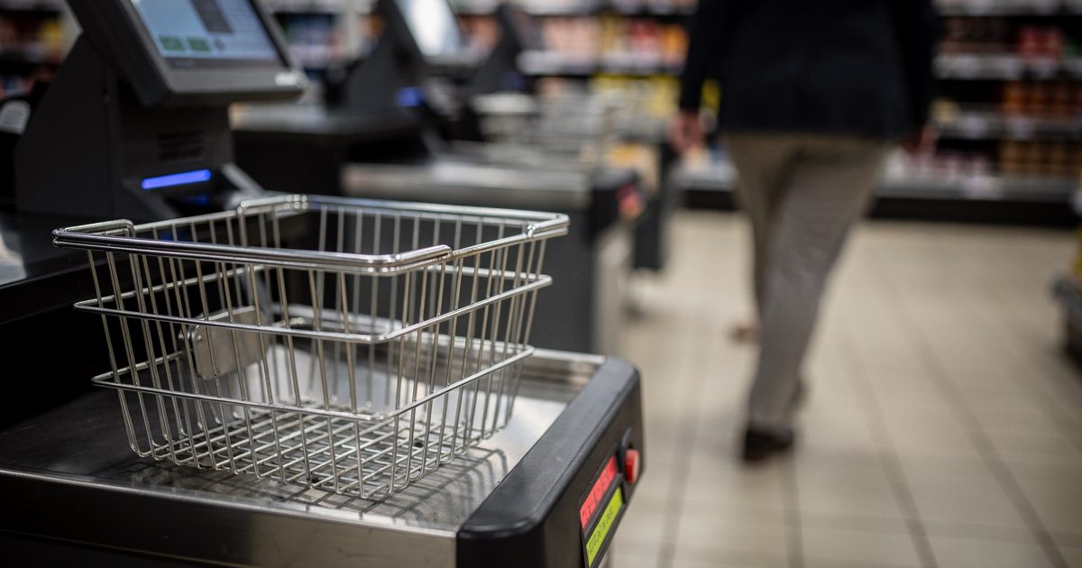 Low-angle shot at the self-checkout of a London supermarket in harsh fluorescent evening light, a shopper visible only from the knees down walking away and.