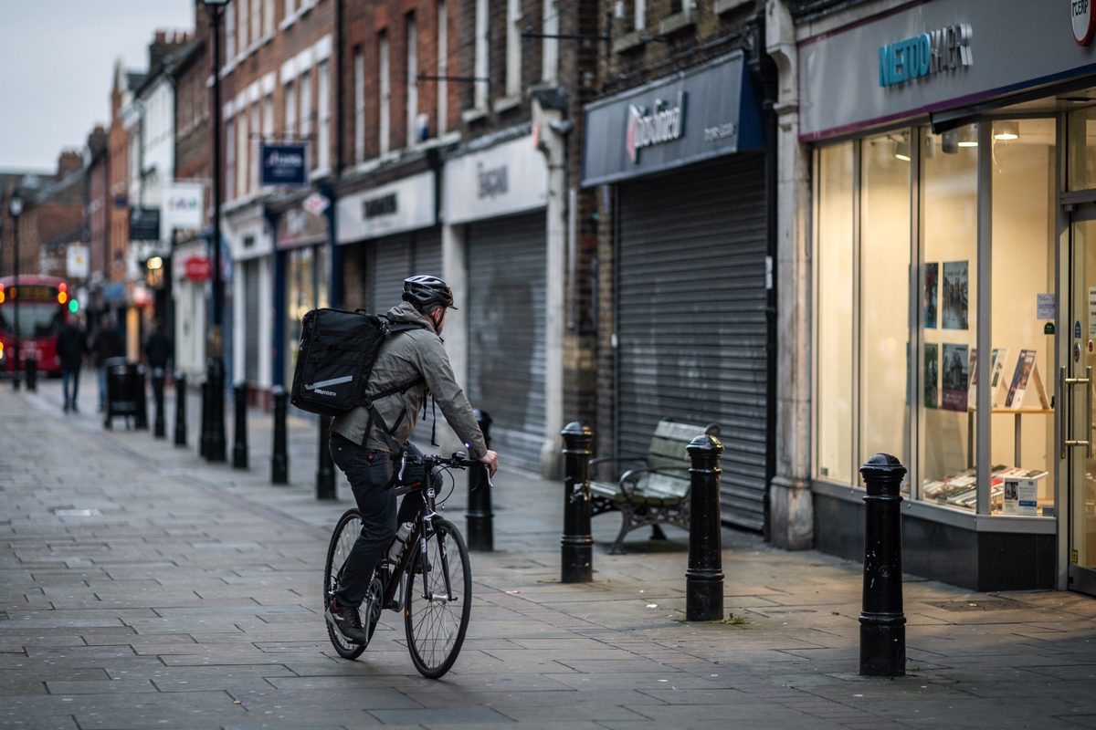 Wide shot in bright, cold midday sun on a pedestrianised English high street, a lone delivery cyclist pedalling past a row of shuttered shopfronts with one.