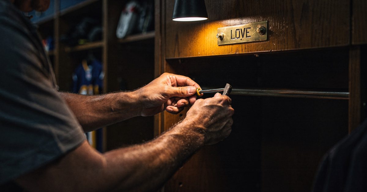 Close shot in a dim NFL locker room lit by a single overhead spot as an equipment manager's forearms, cropped at the elbows, work a flathead screwdriver under.