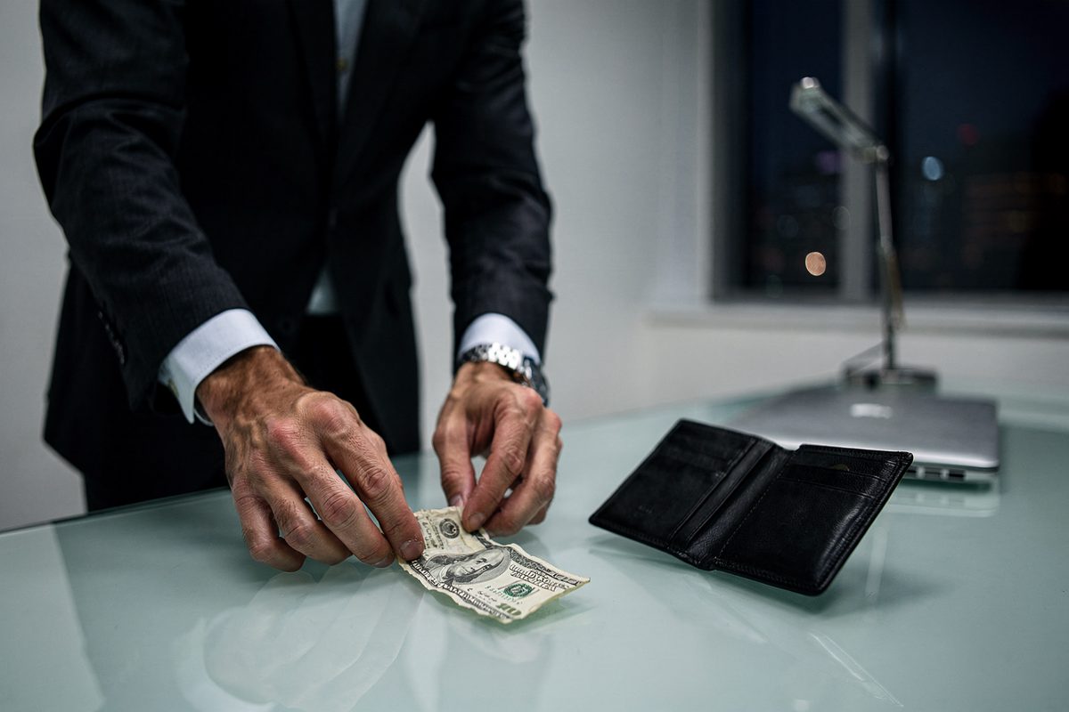 Harsh fluorescent light in a minimalist corporate office as an executive framed from the chest down smooths a crumpled dollar bill onto a glass desk next to an.