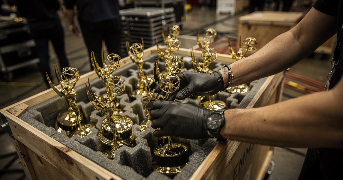 Close shot under warm tungsten backstage work lights of gloved hands cropped at the wrists nesting several winged golden statuettes into foam inside a wooden.