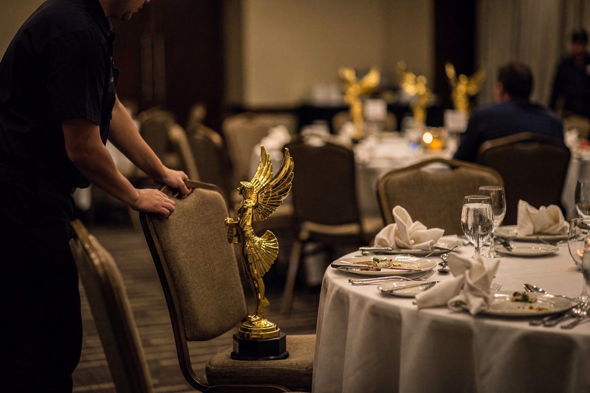 Wide shot in a dim hotel banquet hall after midnight, a golden winged statuette upright in a dining chair beside crumpled napkins and a water glass, a server.