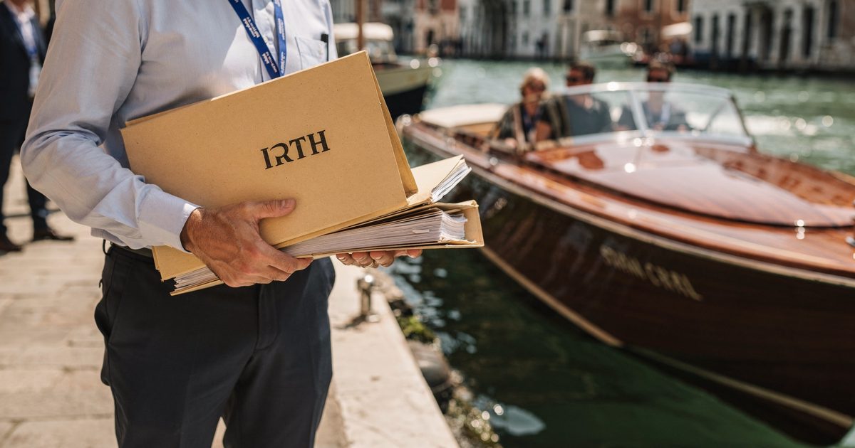 Midday sunlight reflecting off the Grand Canal in Venice, a festival worker framed from the waist down carrying a stack of thick legal folders stamped with.