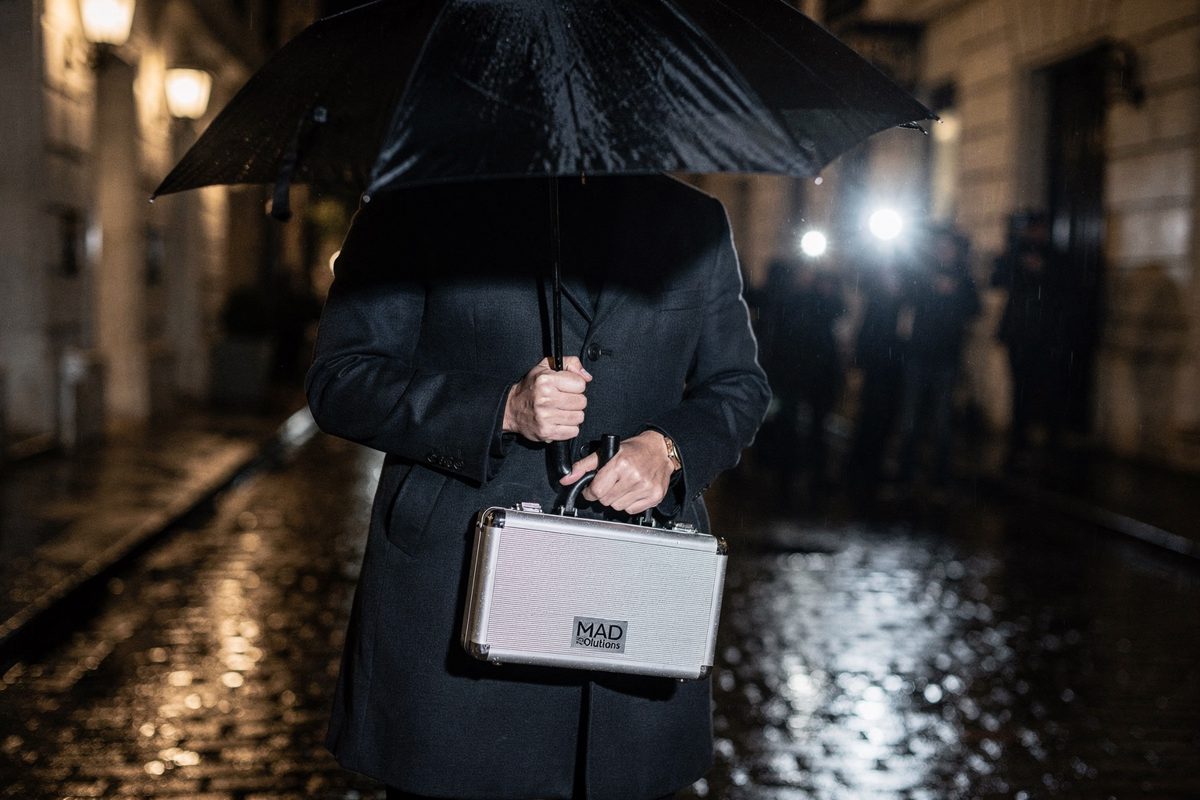 Harsh camera flashes illuminating a rain-slicked cobblestone alleyway at night, a publicist framed from the shoulders down holding a black umbrella over a.