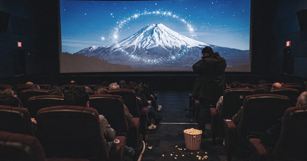 Wide shot from the back row of a half-empty multiplex auditorium at a weekday afternoon showing, cold blue light from a screen filled with a snow-capped.