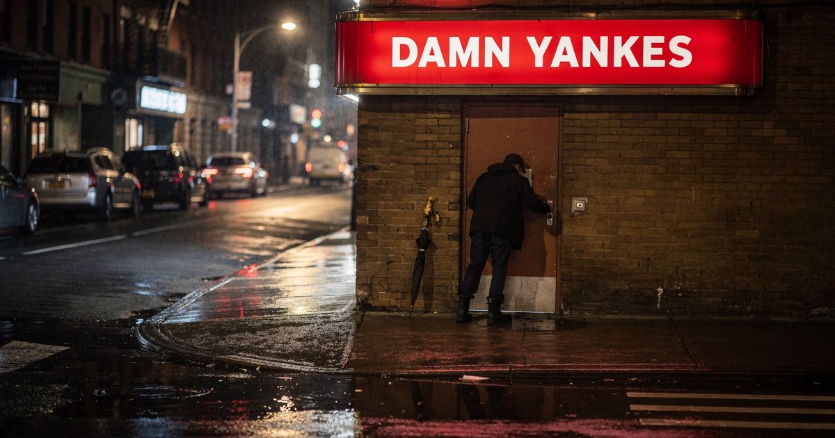 Medium shot from across a wet Manhattan side street at night, a theatre marquee reading "DAMN YANKEES" glowing red above a stage door, a stagehand in black.