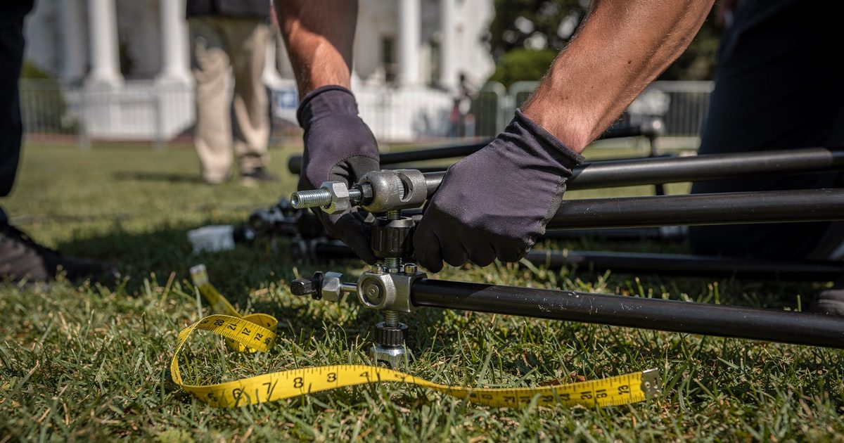 Close detail at waist height on the White House North Lawn in hard early-afternoon sun, a technician's gloved hands cropped at the wrist unscrewing one leg.