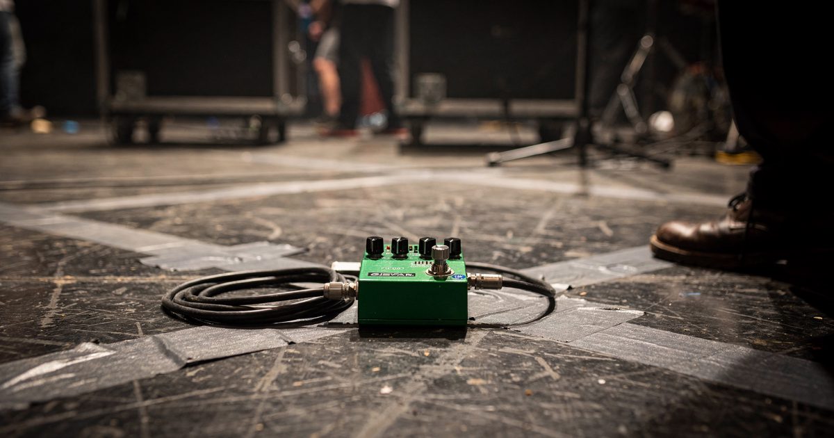 Low close shot at stage-floor level under bare white work lights of a single small guitar loop pedal centred on gaffer-taped boards with its cable coiled.