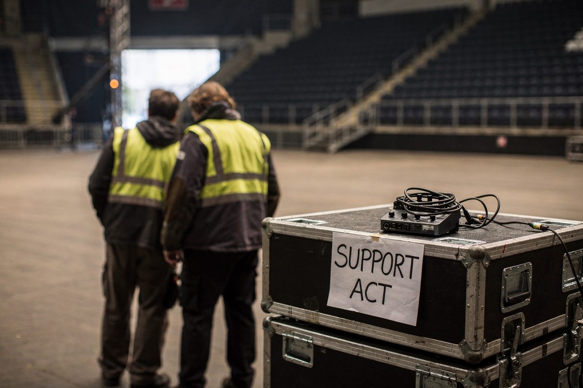 Wide shot across an empty arena floor in mid-afternoon light through open loading doors, two crew in high-vis jackets seen from behind beside a stack of flight.