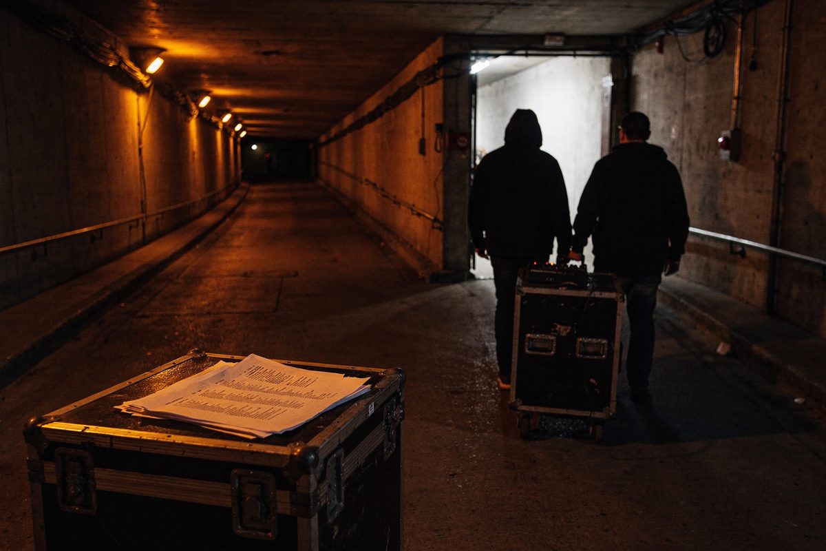 Wide shot down a concrete stadium loading tunnel under orange sodium lamps at night, two crew members in silhouette wheeling a single small black road case.