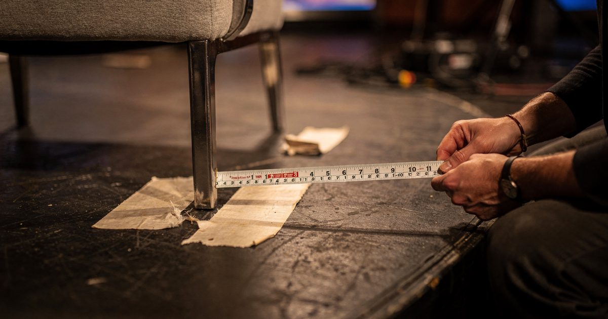 Tight overhead detail under warm tungsten studio lamps of a talk show guest chair leg standing beside an old strip of gaffer tape pressed into the floor, a.