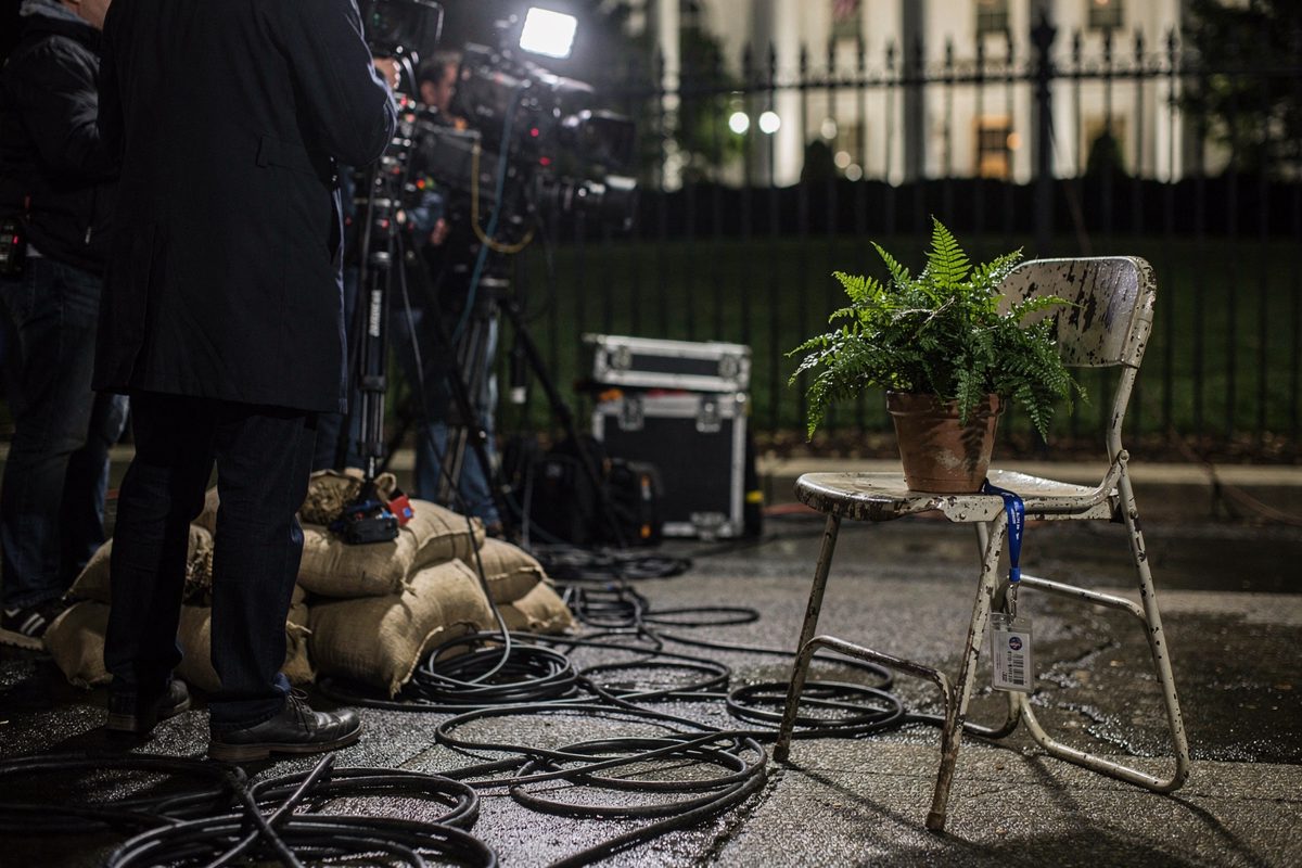 Wide shot under night floodlights along the North Lawn driveway fence, camera operators seen from the waist down among sandbags and cable runs, with one unused.