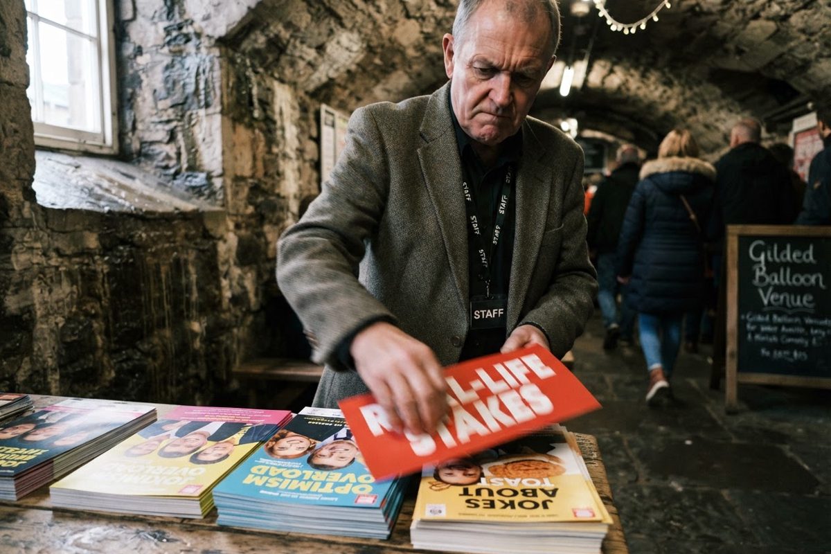 A severe-looking older festival official in a damp cellar, angrily slapping a large red sticker that reads 'REAL-LIFE STAKES' onto a stack of vibrant comedy.