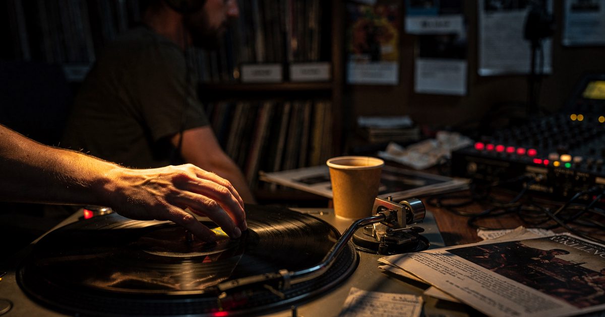 Close detail at night in a small radio studio, a single warm desk lamp catching a hand cropped at the wrist as it lowers a turntable stylus onto a spinning.