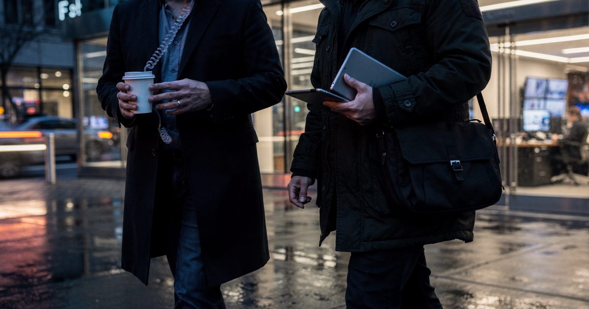 Blue hour outside a midtown network bureau, two producers framed from the chest down crossing a sidewalk toward a glass lobby, one twisting a coiled IFB.