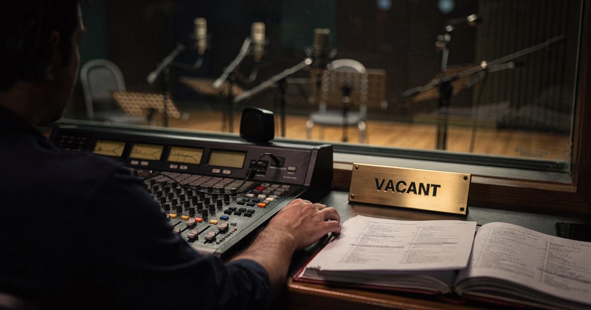 Mid-distance late-afternoon light inside a dubbing stage seen through control-room glass, a sound engineer cropped at the shoulders reaching for a fader while.