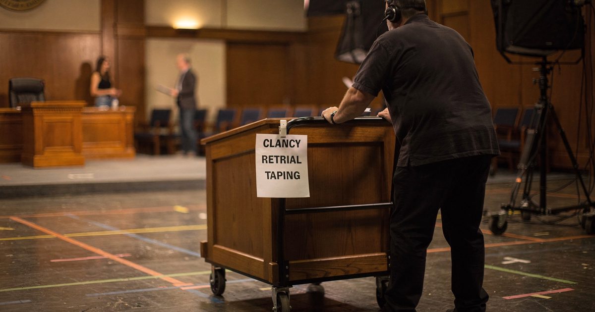 Mid-shot in warm tungsten inside a television courtroom set, a stagehand seen from behind rolling a small wooden jury-box replica across spiked floor marks, a.