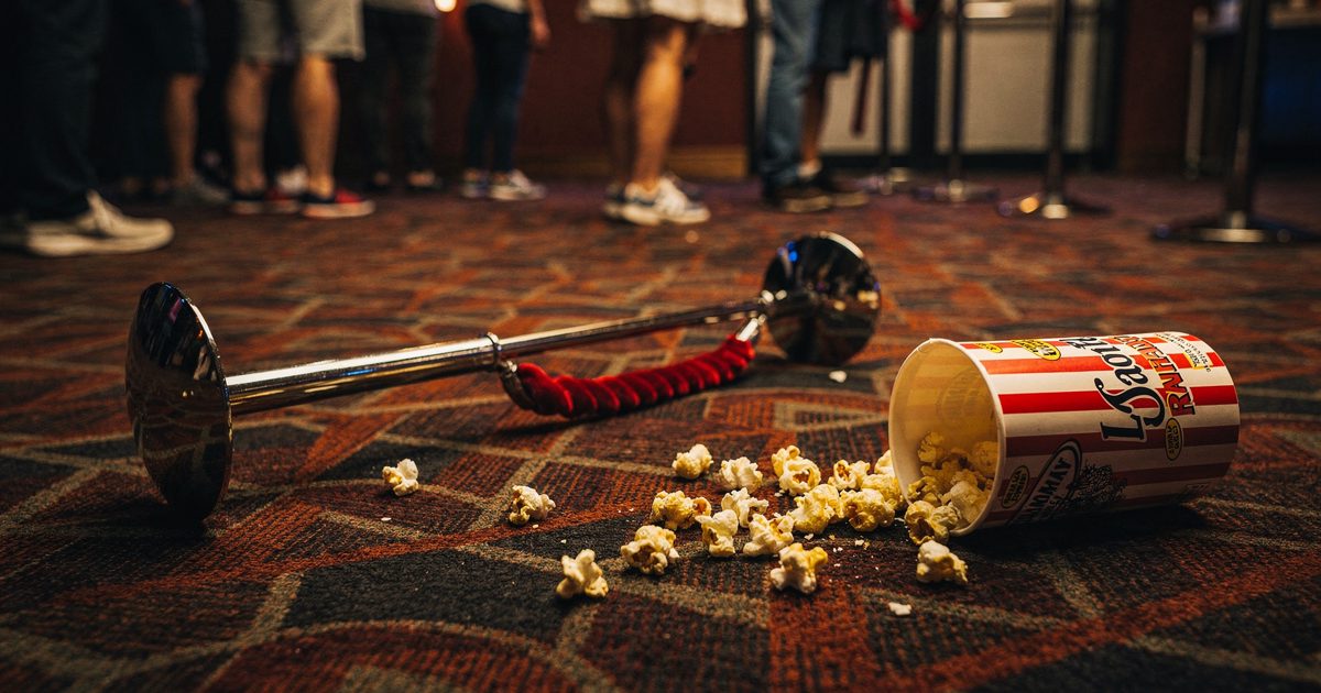 Close detail of a cinema foyer carpet under garish neon lighting, a single velvet rope stanchion knocked sideways while a dropped bucket of popcorn spills.