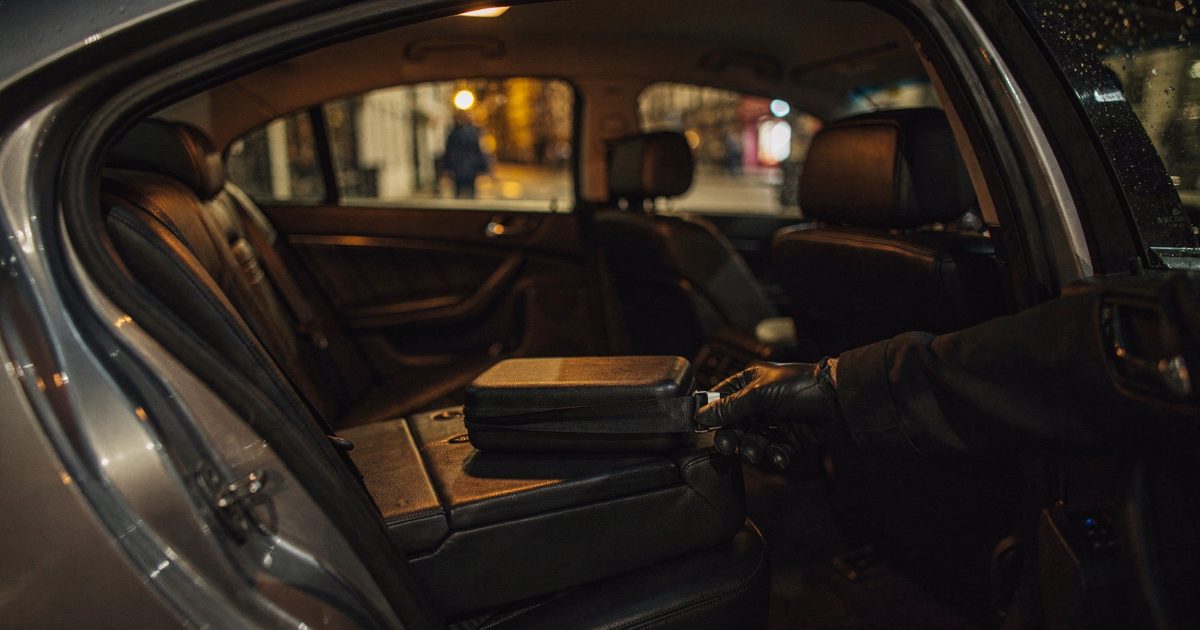 Close shot at night through the open rear door of a grey saloon parked on a narrow Soho street, a gloved hand drawing a seatbelt across a small padded.