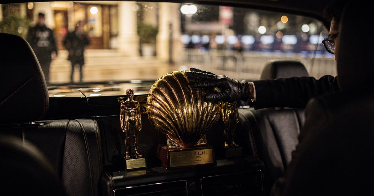 Close detail in warm late-night tungsten glow in the back seat of a festival car idling outside a seafront hotel in San Sebastián, a driver's gloved hand.